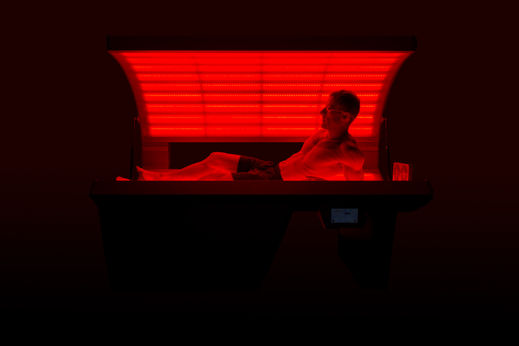 A man lies in a tanning bed, illuminated by red light, undergoing TX red light therapy treatment.